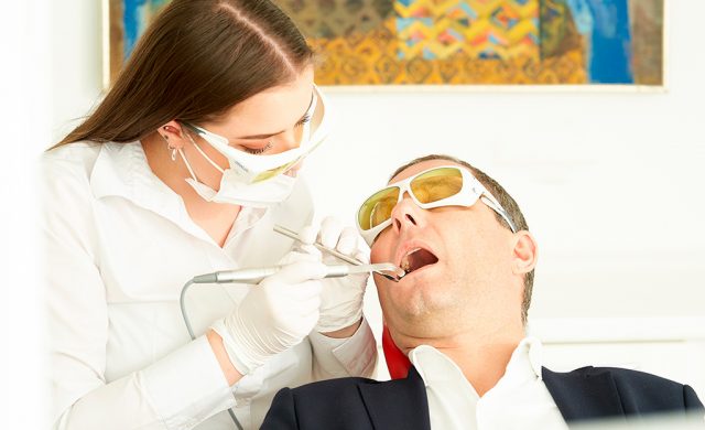 Ein Zahnarzt untersucht einen männlichen Patienten, beide tragen Schutzbrillen; der Zahnarzt benutzt Werkzeuge. Patient sitzt auf einem Stuhl, dahinter bunte Malerei.