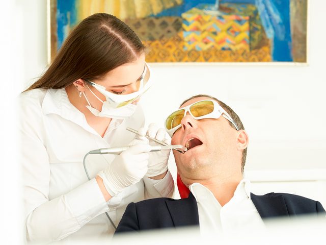 Ein Zahnarzt untersucht einen männlichen Patienten, beide tragen Schutzbrillen; der Zahnarzt benutzt Werkzeuge. Patient sitzt auf einem Stuhl, dahinter bunte Malerei.