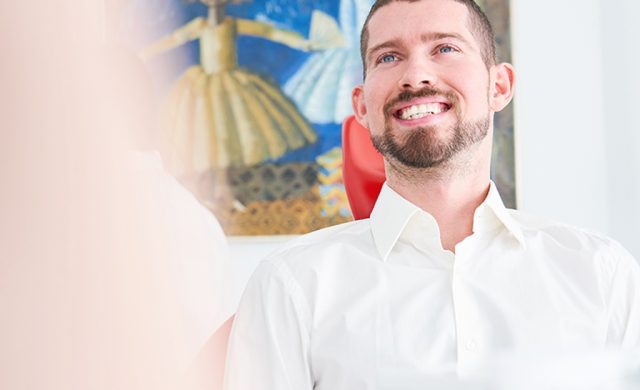Ein lächelnder, bärtiger Mann im weißen Hemd sitzt in einem Haus mit einem bunten Gemälde im hellen, fröhlichen Hintergrund.