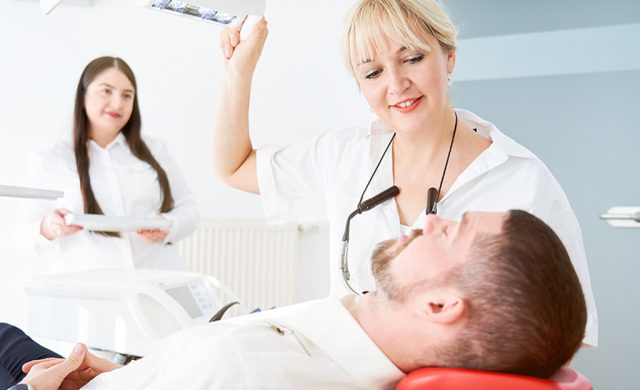 Der Zahnarzt stellt das Licht über dem männlichen Patienten im Stuhl ein; die Assistentin steht dahinter und hält ein Tablett mit Implantatwerkzeugen.