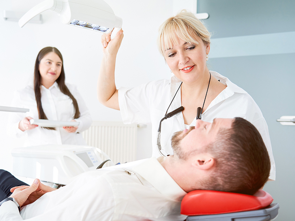 Der Zahnarzt stellt das Licht über dem männlichen Patienten im Stuhl ein; die Assistentin steht dahinter und hält ein Tablett mit Implantatwerkzeugen.