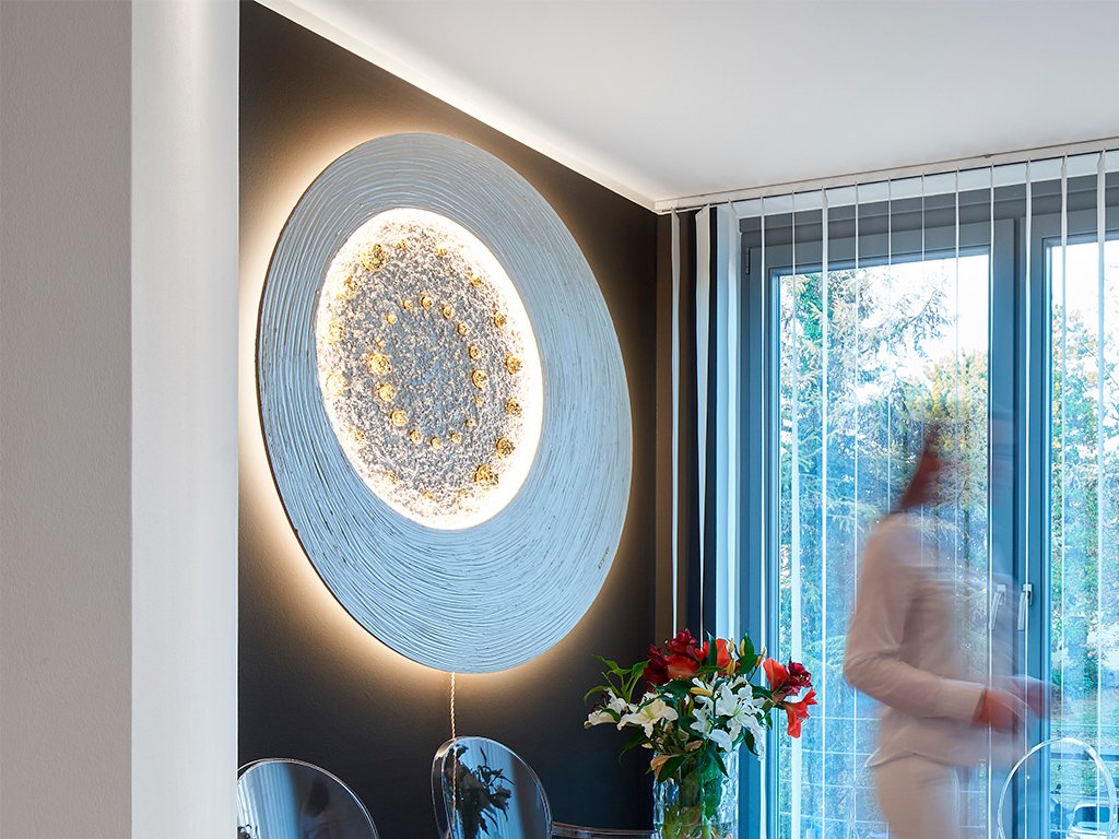 Großes kreisförmiges beleuchtetes Wandgemälde mit konzentrischen Kreisen auf einer schwarzen Wand; eine verschwommene Person geht vorbei, Blumen am Fenster.