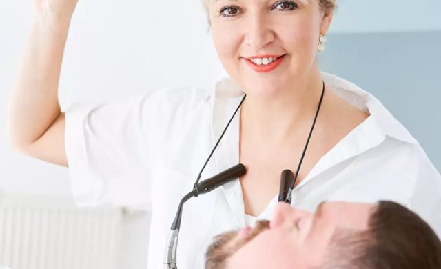 Ein lächelnder Zahnarzt Bad Honnef mit zahnärztlichem Werkzeug steht über einem männlichen Patienten auf einem Stuhl, beide schauen in die Kamera.