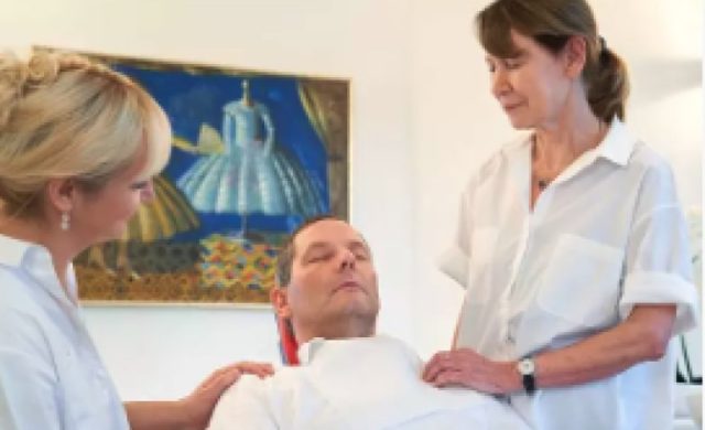 Zwei Frauen in weißen Hemden trösten einen Mann, der am Boden liegt. Hinter ihnen hängt ein buntes Gemälde mit Kleidern an der Wand.