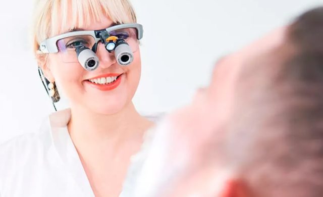 Lächelnder Zahnarzt mit Vergrößerungslupen betrachtet den Patienten im Zahnarztstuhl, weißer Hintergrund.