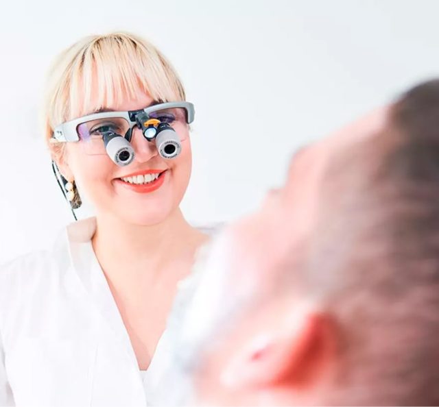 Lächelnder Zahnarzt mit Vergrößerungslupen betrachtet den Patienten im Zahnarztstuhl, weißer Hintergrund.